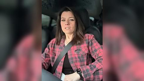 Media: Video of a woman in a car, wearing a red plaid shirt and black top, with a grey seatbelt, seated in a black car seat, blurred background.