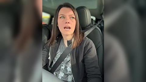 Media: Video of a woman with straight brown hair, wearing a gray patterned blouse, and a black jacket, sitting in a car with a seatbelt on, looking surprised or concerned, blurred background.