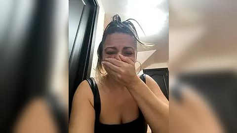 Media: A candid video of a woman with light skin and brown hair tied back, wearing a black top, covering her mouth with her hand, in a bathroom with a mirror and white light.