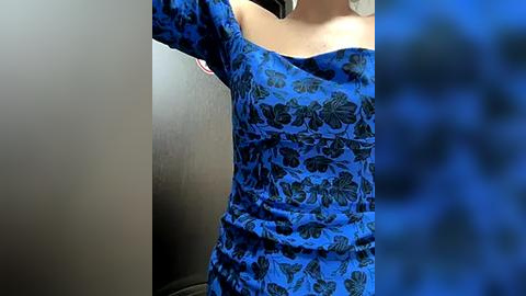 Media: Video of a woman's upper body in a blue floral dress, with a blurred blue background and a black smartphone in her right hand.