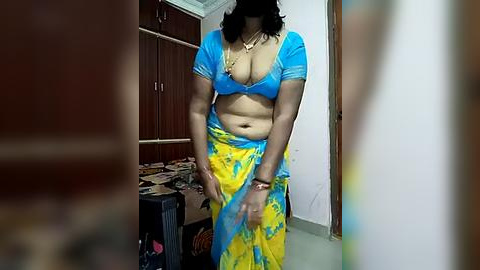 Media: Video of a curvy, dark-skinned woman in a blue and yellow sari, standing in a cluttered room with wooden cabinets and a messy desk.