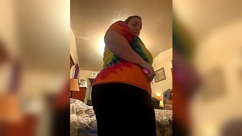 Media: Video of a plus-size woman in a colorful tie-dye top and black pants, adjusting her bra in a dimly lit bedroom with a bed, framed artwork, and a TV visible.