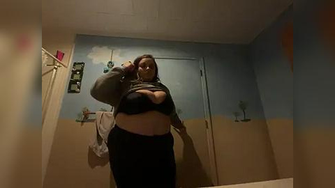 Media: A video of a plus-size woman with dark skin, short brown hair, wearing a black bra and high-waisted pants, taking a selfie in a beige-walled bathroom with a blue sky mural and a door.