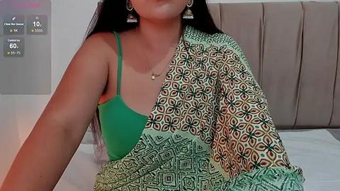 Media: Video of a South Asian woman with long black hair, wearing a green bra and a multi-colored patterned sari, sitting on a bed with a beige headboard.