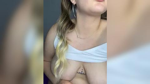 Media: Video of a woman with light skin and long, wavy blonde hair, wearing a white off-the-shoulder top, exposing her large breasts with a small tattoo between them.