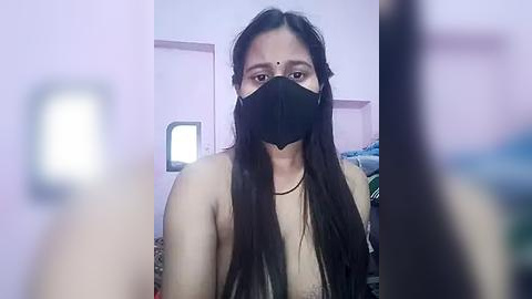 Media: Video of a topless South Asian woman with long black hair, fair skin, and a black mask, standing in a dimly lit, cluttered room with pink walls.