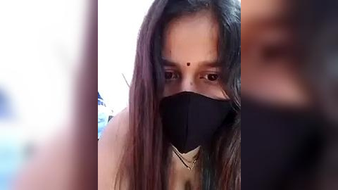 Media: Video of a woman with long dark hair, wearing a black face mask and a beige top, looking towards the camera, blurred background.