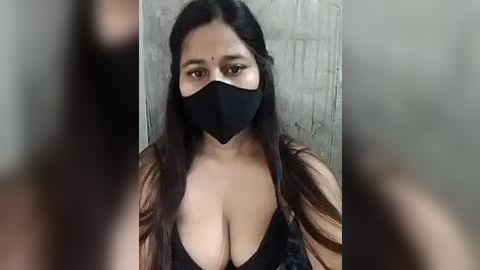 Media: A video of a South Asian woman with long black hair and a black face mask, wearing a low-cut black top revealing ample cleavage. She stands against a textured, light-colored wall.