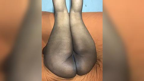 Media: A video of a woman lying on her back, wearing sheer black stockings, with her legs bent and raised, showcasing her buttocks prominently. The background features a light blue wall and an orange bedspread.