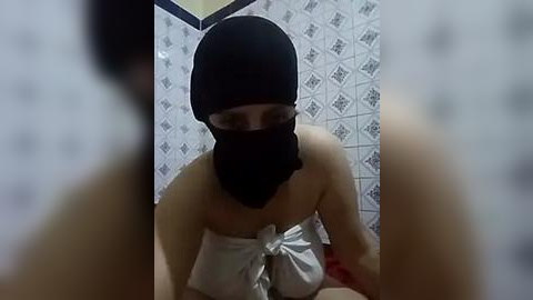 Media: Video of a woman in a black balaclava and white towel, seated in a tiled bathroom with geometric patterned walls. The image is slightly blurred in the foreground.