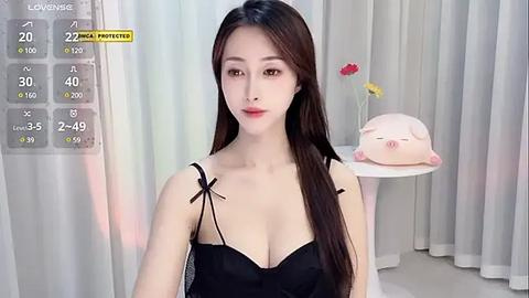 Media: A video of an East Asian woman with long, straight black hair, fair skin, and wearing a black lingerie top. She stands in front of a white background with a pink piglet figurine on a white table.