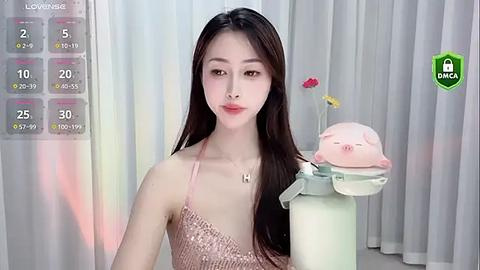 Media: Video of a young Asian woman with long black hair, wearing a pink sequined dress, holding a plush toy. Background includes a virtual game interface with health and stamina bars, a green shield icon, and a pink flower.