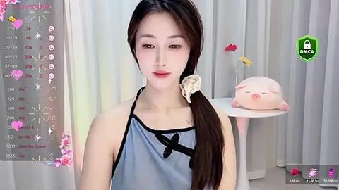 Media: A video of a young East Asian woman with fair skin, straight black hair in a ponytail, wearing a blue apron. The background features a white curtain and a pink pig plush toy on a table.