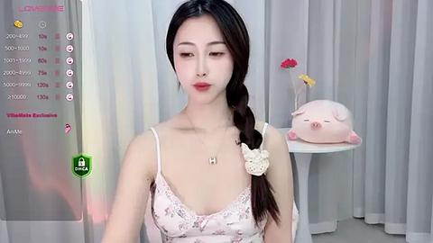 Media: Video of an Asian woman with long braided hair in a pastel pink camisole, wearing a scrunchie, standing in a room with a pig-shaped pillow.