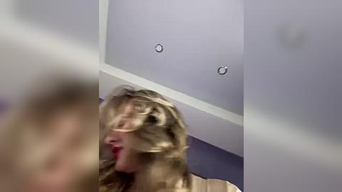 Media: Video of a blonde woman with messy hair and red lipstick, seated at a table in a dimly lit room with a lavender ceiling and a white wall.