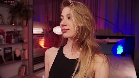 Media: Video of a slender, blonde woman with long hair, wearing a black tank top, standing indoors with a potted plant, shelves, and a bed with blue lights in the background.