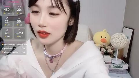 Media: Video of an East Asian woman with short black hair, wearing a white top and pink accessories, in a room with a yellow duck and flowers, overlaid with social media stats.