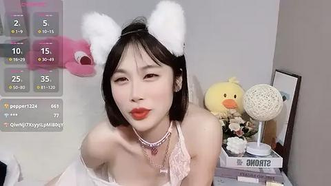 Media: Video of an East Asian woman with fair skin, wearing a white bunny headband, pink lingerie, and red lipstick, standing in a bedroom with a white bed, yellow stuffed duck, and pink flowers.