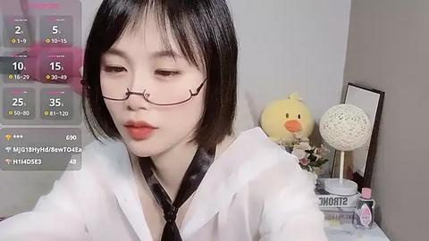 Media: Video of an Asian woman with straight black hair and glasses, wearing a white shirt, in a room with a yellow duck, books, and a clock.