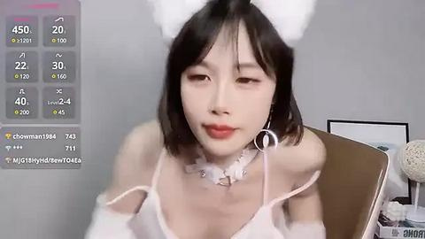 Media: Video of an East Asian woman with short black hair and white bunny ears, wearing a white off-shoulder top and a silver necklace. She sits in front of a gray background with a digital camera display.