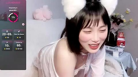 Media: Video of an East Asian woman in a white nightgown with cat ears, smiling, in a cozy room with plush toys and a laptop screen showing stats.