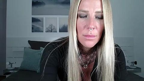 Media: Video of a blonde woman with multiple facial piercings, wearing a black top, in a modern bedroom with minimalist decor.