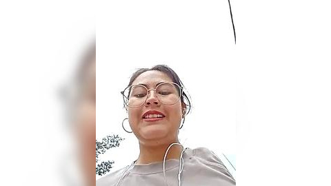 Media: Video of a happy, light-skinned Asian woman with brown hair in glasses, wearing a light gray top, and white earphones, looking upward. Background is bright and blurry.