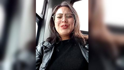 Media: Video of a smiling, light-skinned woman with glasses, wearing a black leather jacket and a black ribbed top, sitting in a car.