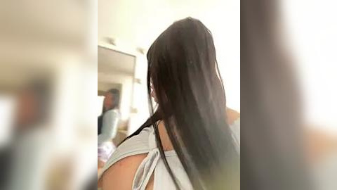 Media: Video of a woman with long, dark hair, seen from behind, in a modern, brightly lit room with a large mirror reflecting her blurred reflection.