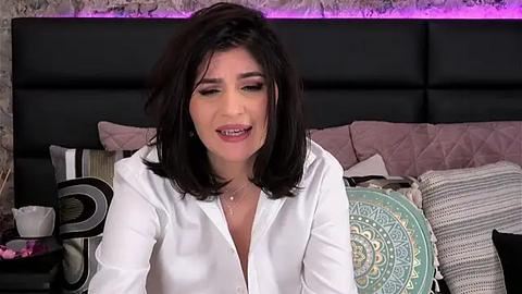 Media: Video of a woman with shoulder-length dark hair, wearing a white button-up shirt, sitting on a bed with a black headboard and pastel-colored pillows, smiling.