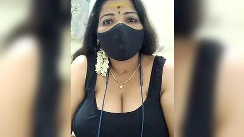 Media: A video of a woman with medium brown skin, wearing a black face mask, floral headband, and a black sleeveless top, featuring large breasts. She has long, straight black hair and is in an indoor setting.