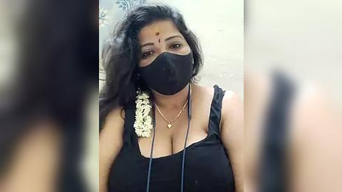 Media: Video of a South Asian woman with medium skin tone, wearing a black mask, black tank top, and a white flower behind her ear. Background is blurred, showing a neutral setting.