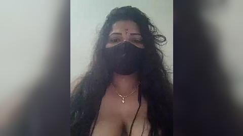 Media: A video of a woman with long, curly black hair and a black face mask, wearing a gold necklace and a red bindi. She has a fair complexion. The background is blurry and indistinct.