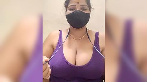 Media: A video of a woman with a medium skin tone, wearing a purple tank top with large cleavage, black face mask, and a gold necklace, indoors, blurred background.