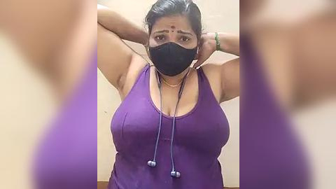 Media: Video of a middle-aged South Asian woman with medium skin tone, wearing a purple tank top, black face mask, and headphones, stretching her arms. Background shows blurred figures in a dimly lit room.