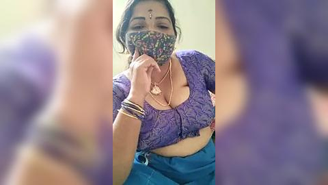 Media: Video of a South Asian woman with medium skin tone, wearing a purple lace top, blue high-waisted skirt, and floral face mask, with multiple bracelets on her wrist.