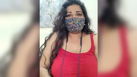 Media: Video of a South Asian woman with long, wavy black hair, wearing a red tank top, floral face mask, and headphones, against a blurred background with floral wallpaper.