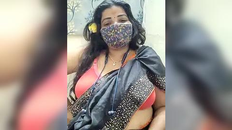 Media: Video of a woman with medium-dark skin, long dark hair, wearing a colorful patterned mask, a pink bra, and a black sari with floral patterns. She appears to be indoors with blurred background.