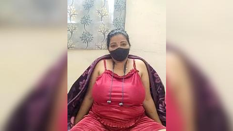 Media: A video of a South Asian woman with medium skin tone, dark hair, and a black face mask, wearing a red sleeveless top and matching pants, seated on a purple chair in a plain room with a floral-patterned curtain in the background.