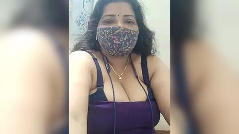 Media: Video of a curvy South Asian woman with long, wavy dark hair, wearing a floral-patterned face mask, a purple top, and a gold necklace, standing in a bathroom with floral tiles.