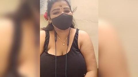 Media: Video of a South Asian woman with medium skin tone, wearing a black face mask, black tank top, and floral hair accessory, standing indoors.