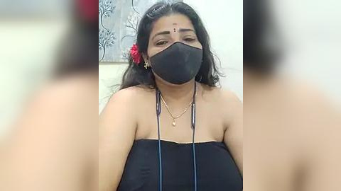 Media: Video of a South Asian woman with medium skin tone, long black hair adorned with a red flower, wearing a black face mask, black strapless top, and gold necklace, against a white and floral-patterned wall background.