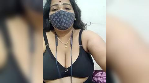 Media: A close-up video of a woman with medium skin tone, wearing a blue face mask, black bra, and necklace, with a blurred background.