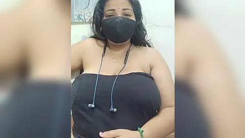 Media: Video of a South Asian woman with medium skin tone and long black hair, wearing a black strapless dress, black face mask, and a gold necklace. Background shows a blurred wall.