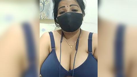 Media: A video of a South Asian woman with a medium complexion, wearing a blue bra, black face mask, and a gold necklace. She has dark hair tied back and is indoors, with blurred background.