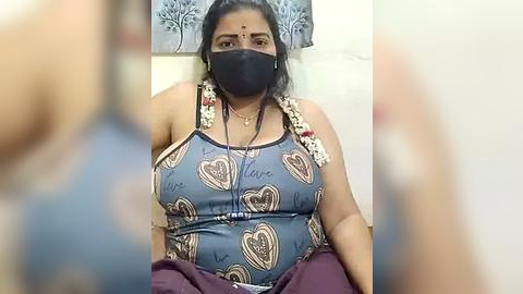 Media: A video of a plus-sized woman with dark skin, wearing a face mask, floral-patterned tank top, and maroon pants, sitting indoors, partially blurred background.