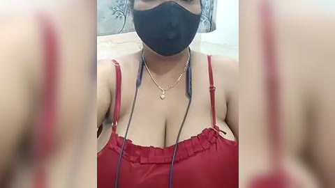 Media: Video of a woman wearing a black face mask, red frilled camisole, and gold necklace, with blurred arms and background.