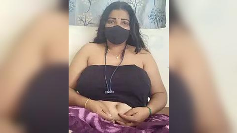 Media: Video of a South Asian woman with medium skin tone, wearing a black face mask, black strapless top, and necklace. Background includes a white headboard and floral-patterned wall.