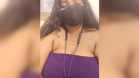 Media: Video of a woman with long dark hair, wearing a purple strapless top, black face mask, gold necklace, and earphones, blurred background.