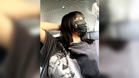 Media: Video of a woman with straight black hair and a black mask, wearing a grey T-shirt with a graphic design. She's in a car, with the blurred background showing a grey textured ceiling and a white object.